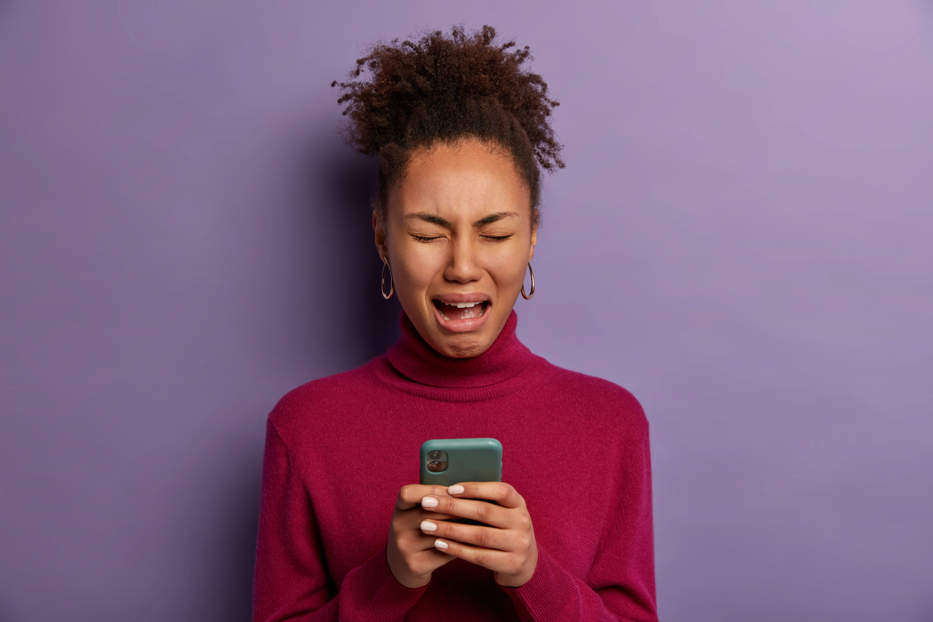 Depressed crying woman holds mobile phone in her hands, wears burgundy turtleneck, upset not to receive message from boyfriend, has no money to call friend, stands with sorrowful expression.